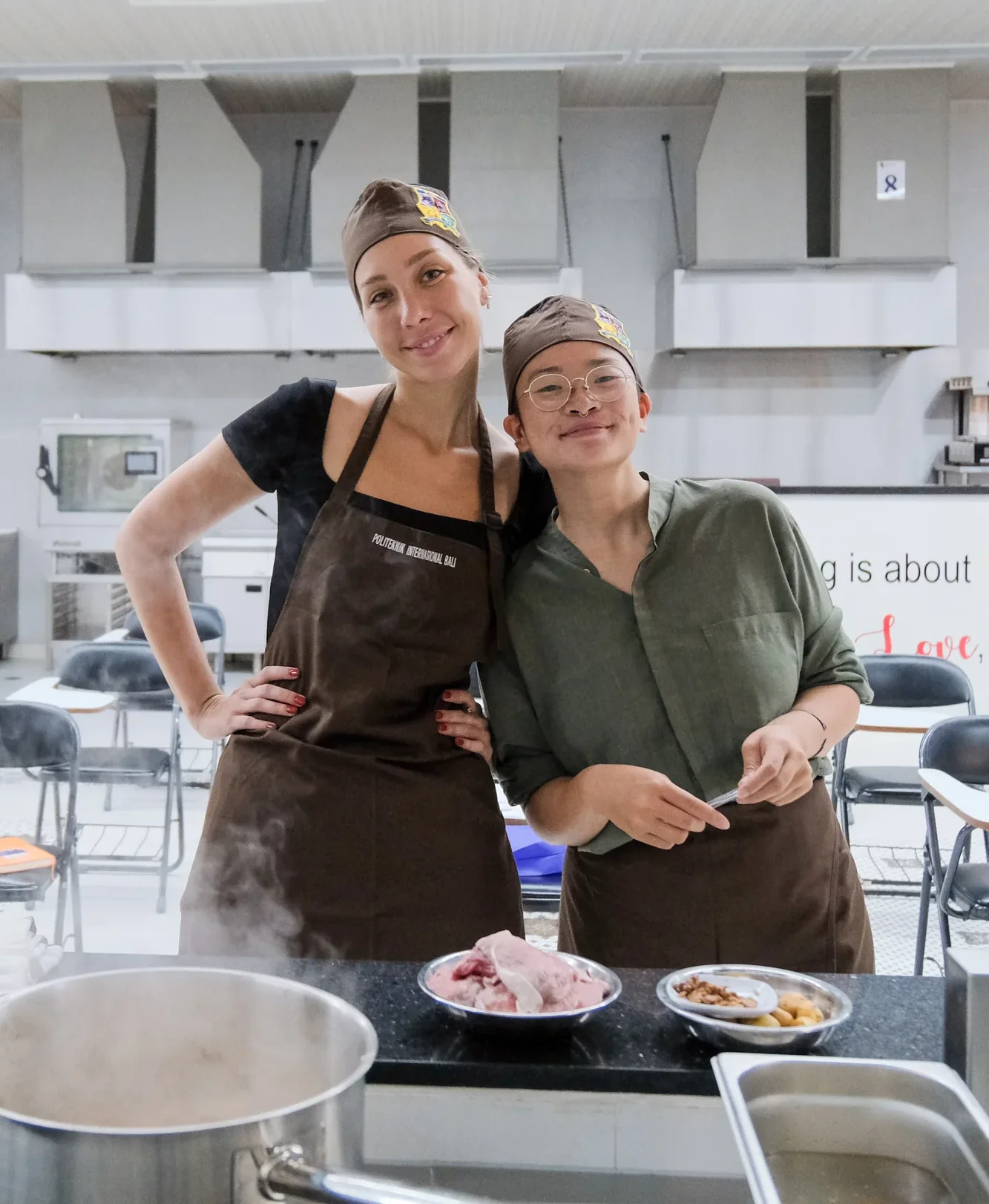 Two culinary students posing in aprons beside pots and prepared ingredientsollege kitchen