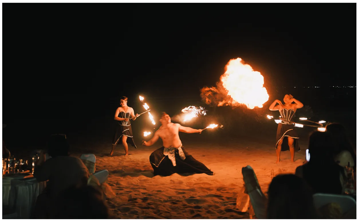 Two fire performers on the beach at night, one breathing a large flame